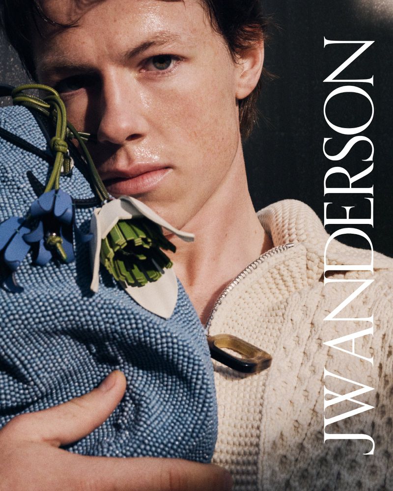 Close-up shot of a light-skinned male model holding a floral-design bag and wearing a white zip-up sweater in studio lighting.