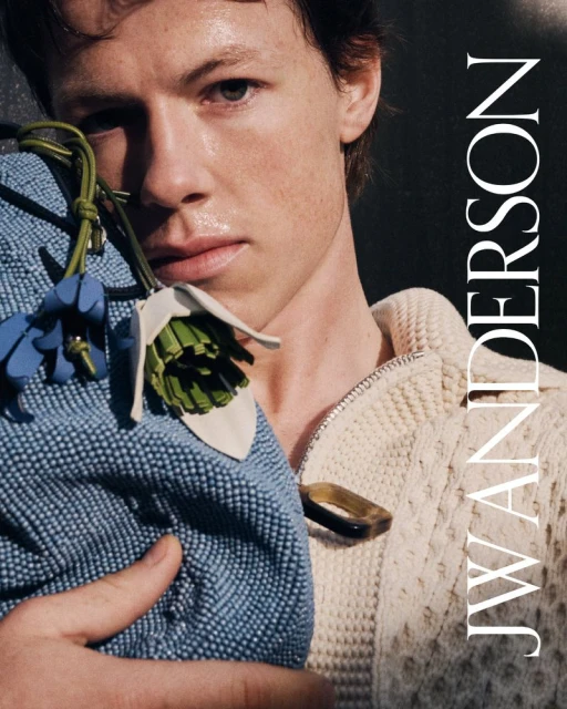 Close-up shot of a light-skinned male model holding a floral-design bag and wearing a white zip-up sweater in studio lighting.
