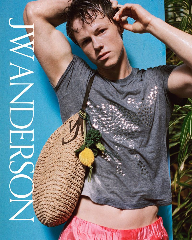 A slender man poses with a woven bag adorned with a lemon, set against a blue wall and green foliage for a fashion editorial.