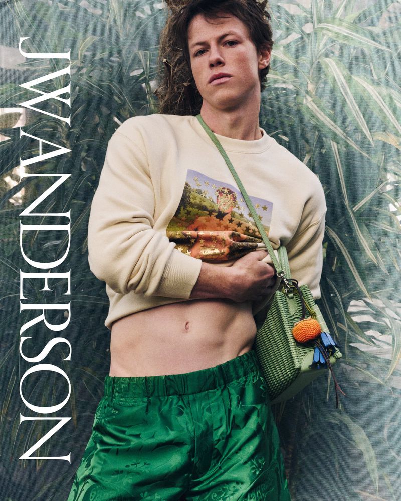 Male model in editorial fashion shot wearing a pale yellow sweatshirt, green shorts and carrying a green purse in a lush outdoor setting.