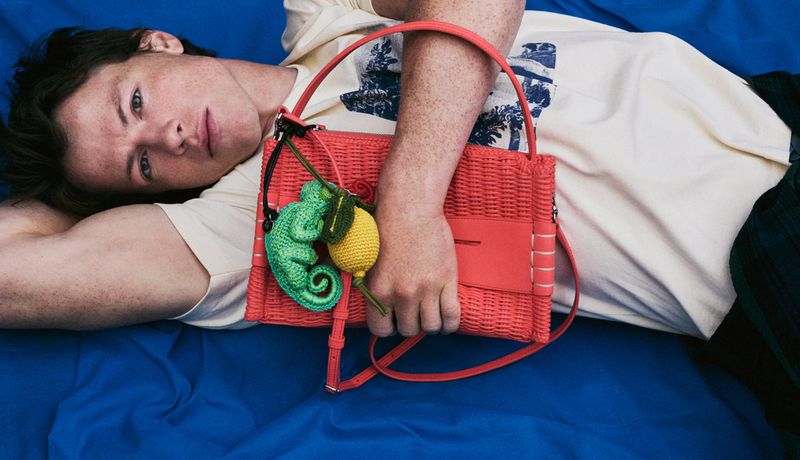 Model lying on vibrant blue fabric holding a bright pink woven handbag with green chameleon and yellow lemon charms.