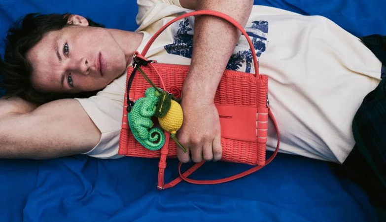 Model lying on vibrant blue fabric holding a bright pink woven handbag with green chameleon and yellow lemon charms.