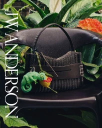 J.W. Anderson dark woven handbag with a crochet chameleon charm resting on a garden chair among lush green leaves.