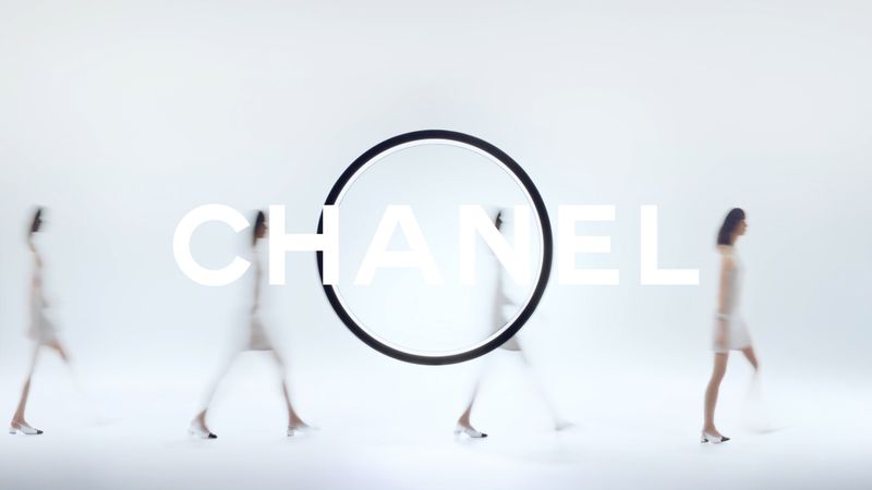 Fashion models walk against a bright white backdrop with a Chanel logo and a black circular frame in a bright and airy studio.