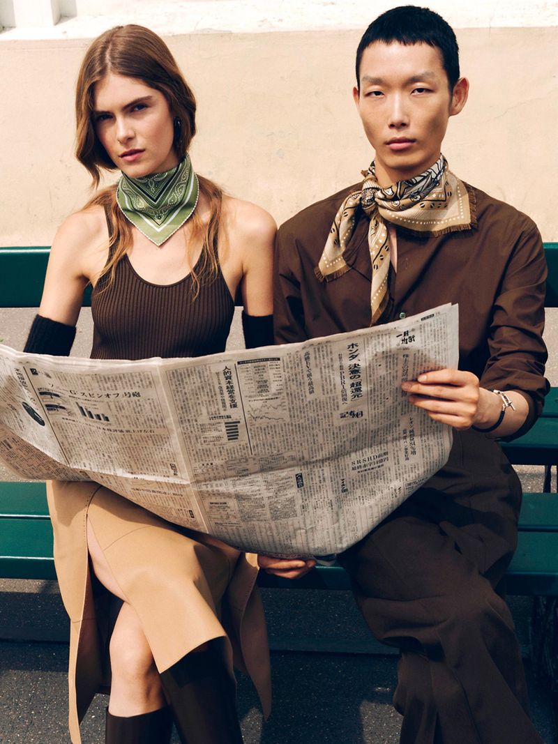 Stylish couple reads newspaper on green bench. Young Asian man in brown shirt, Caucasian woman in tank top, both wear neck scarves.