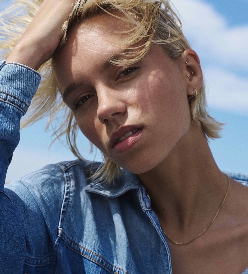 Close-up portrait of an adult female with short blonde hair, wearing a denim jacket against a clear sky backdrop in natural lighting.