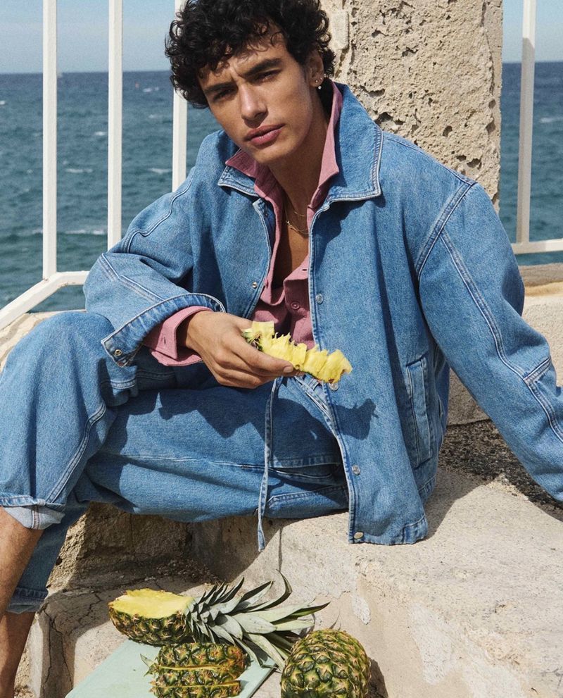 Young adult male sits on stone steps near the ocean and eats sliced pineapple; wearing denim jacket, pink shirt, and denim pants.