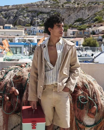 A man in a beige jacket and shorts stands on a sunny dock with a fishing net and toolbox in an outdoor, daytime shot.