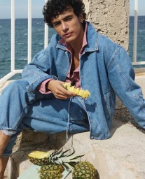 Man in a blue denim jacket and matching jeans holding a slice of fresh pineapple on a stone ledge overlooking the sea.
