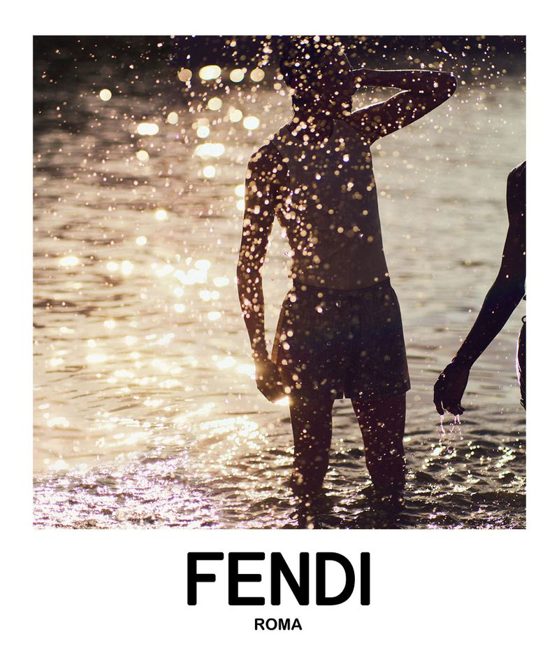 Two people are partially submerged in shallow, sunlit water at dusk. The sunlight causes a shimmering effect on the water surface.