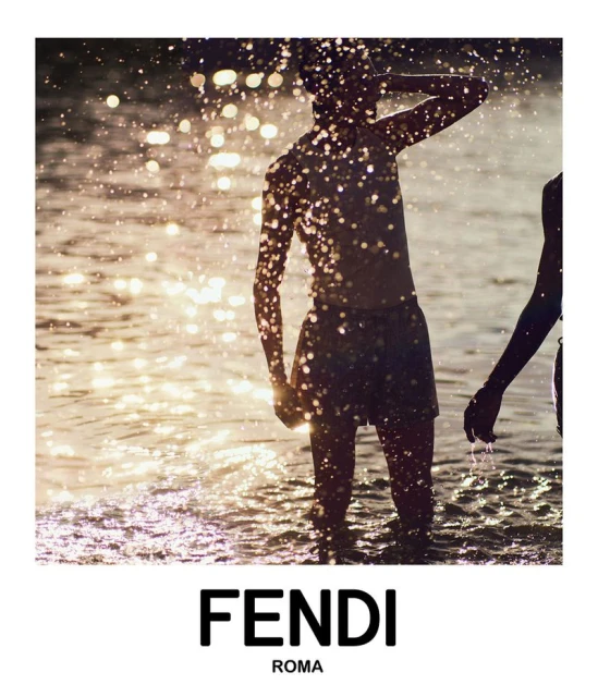 Two people are partially submerged in shallow, sunlit water at dusk. The sunlight causes a shimmering effect on the water surface.