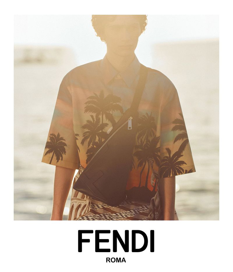 Person wearing a palm tree patterned shirt and black crossbody bag stands in front of a sunlit body of water for Fendi advertisement.
