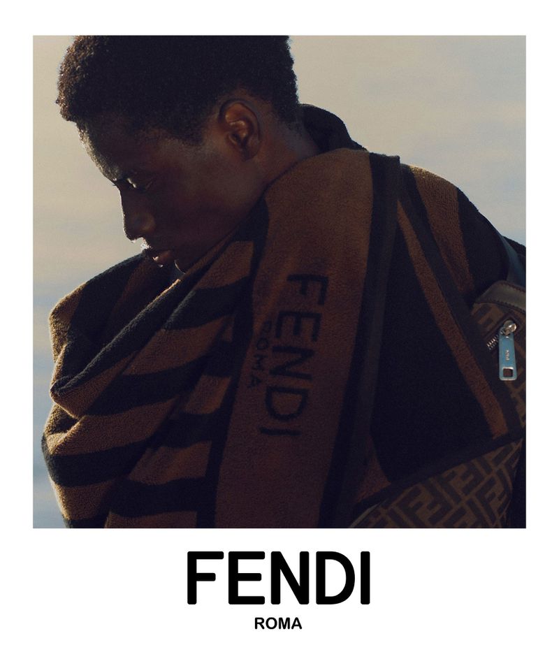 Male model poses with a brown Fendi towel draped over his shoulder, bag with a signature pattern, and the Fendi logo in the foreground.