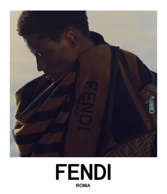 Male model poses with a brown Fendi towel draped over his shoulder, bag with a signature pattern, and the Fendi logo in the foreground.