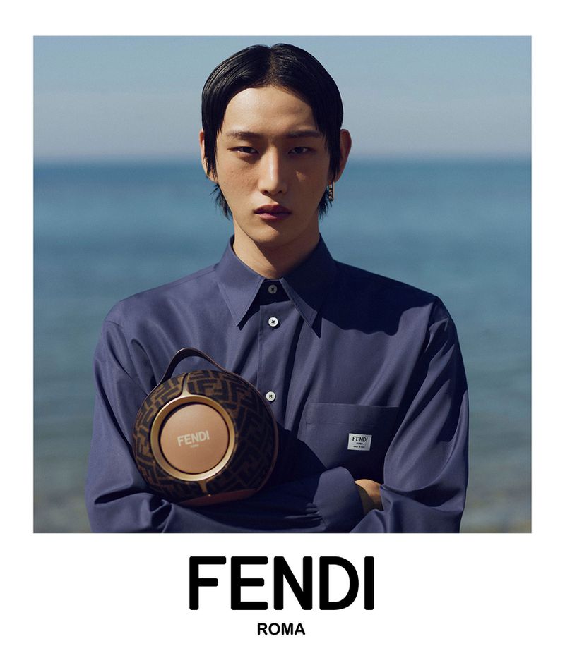 A young adult male poses in a medium shot wearing a blue collared shirt, holding a round patterned bag with the Fendi logo near an ocean backdrop.