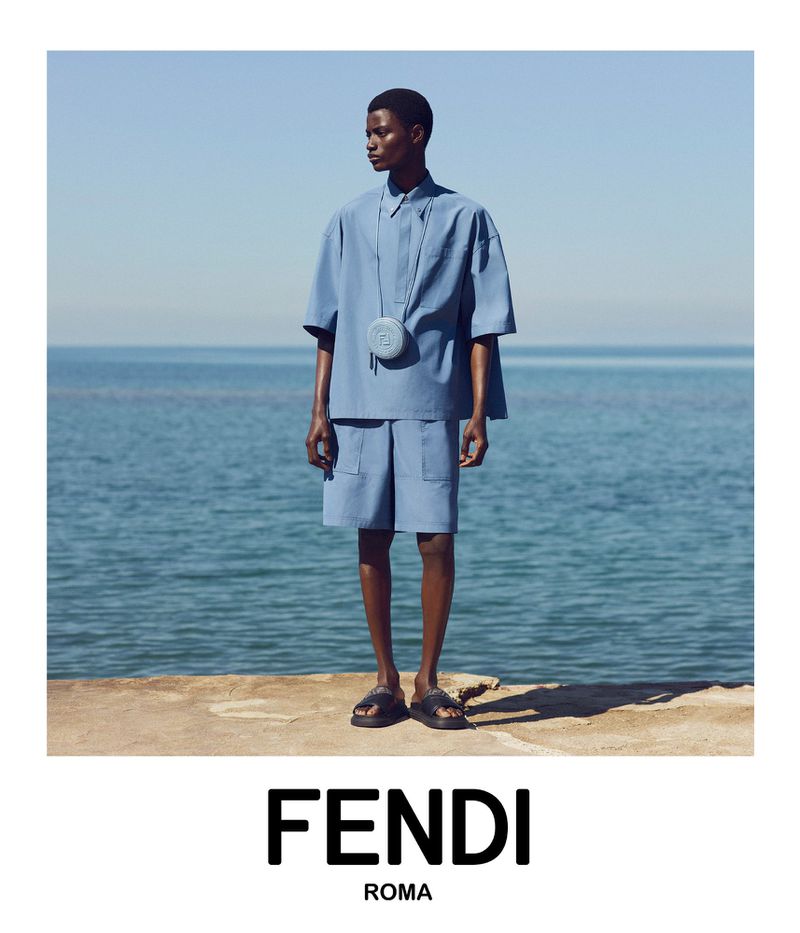 A dark-skinned male model wears a blue shirt and shorts outfit standing in front of a sea backdrop on a stone structure with Fendi Roma text.