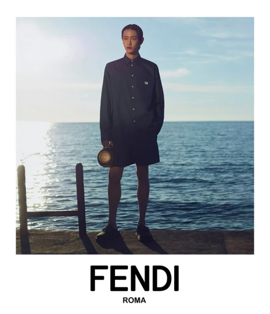 Male fashion model in long-sleeved shirt and shorts standing on a platform with a round bag, with ocean and sky backdrop.