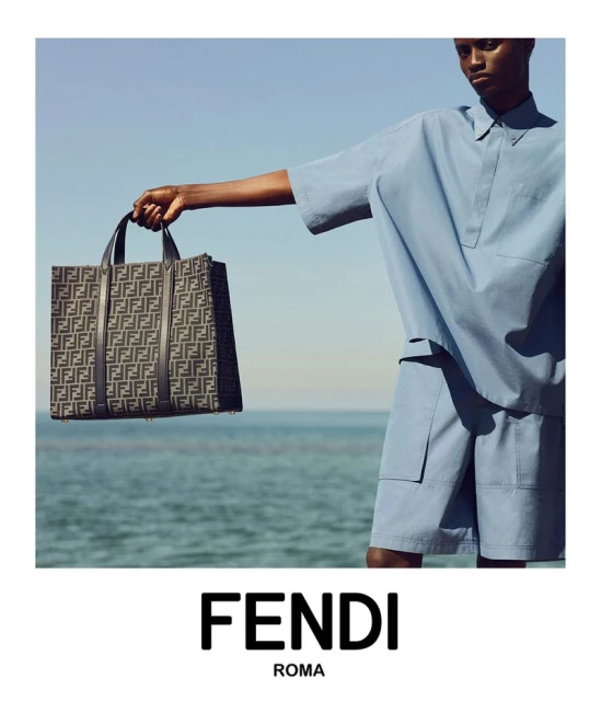 An adult Black male presents a patterned Fendi tote bag on an ocean beach in bright daylight wearing a matching blue outfit.