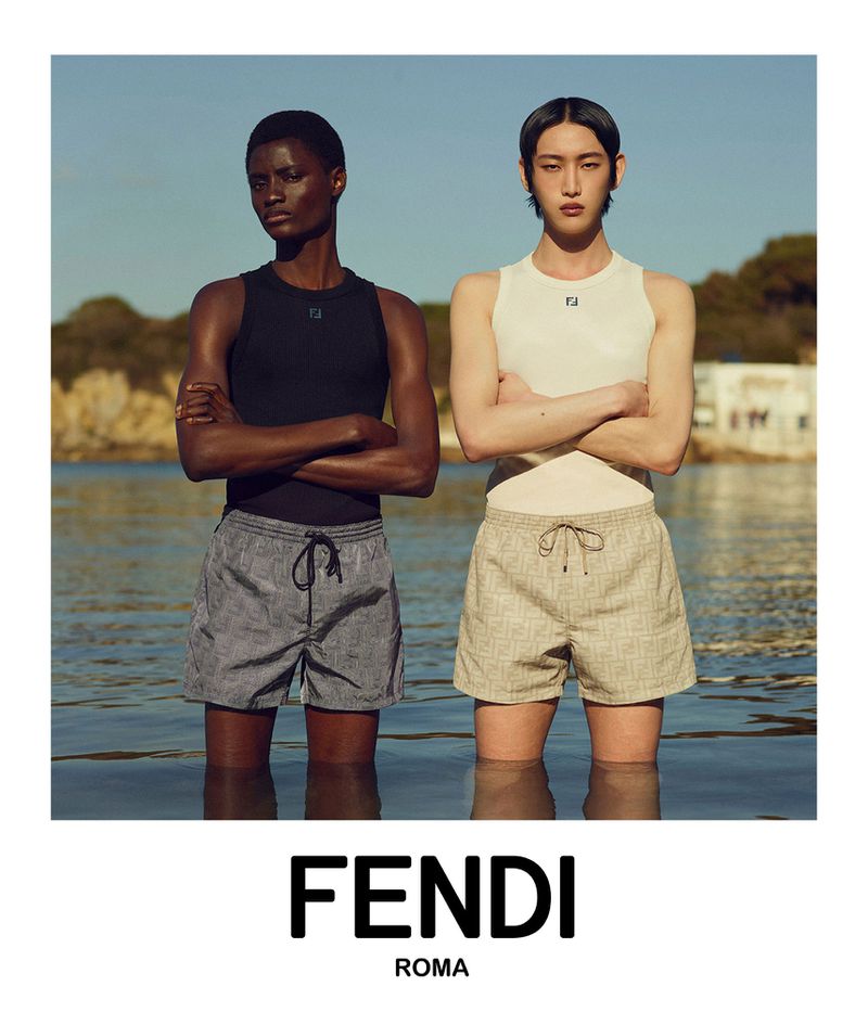 Two people are standing waist-deep in the ocean wearing Fendi branded swimsuits. The sky is blue and clear in the background.
