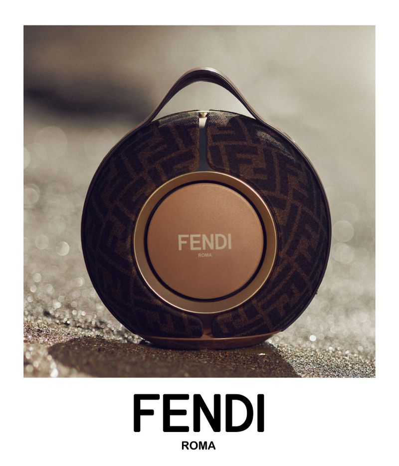 Fendi branded spherical speaker resting on a sandy surface. Speaker has a leather handle, brown FF logo pattern, and a bronze center.
