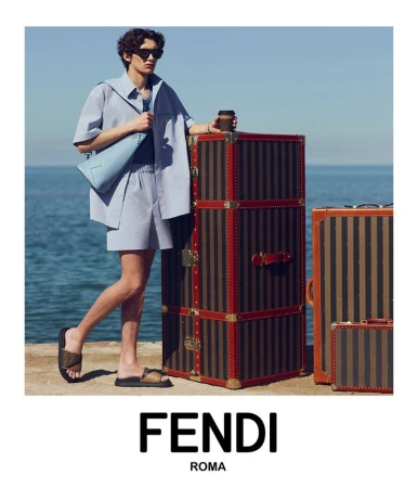 Man in a light blue outfit standing next to stacked brown suitcases with a view of the ocean and a light blue sky in the background.