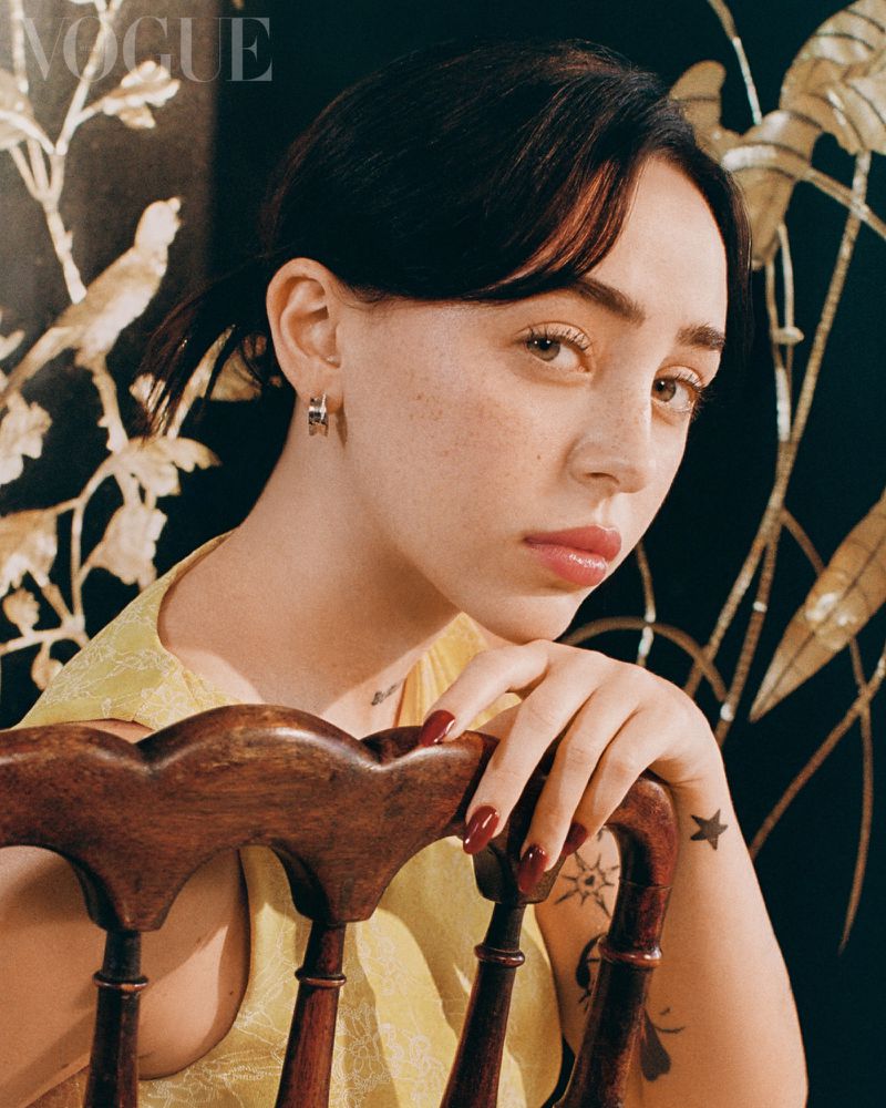 Close-up portrait of a young adult female with dark hair and light skin, wearing a yellow top and earrings, sitting on a wooden chair.