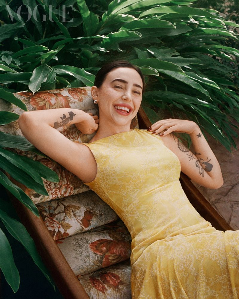 Smiling woman in yellow lace dress lounging on floral couch surrounded by lush green foliage in an outdoor, natural light setting.