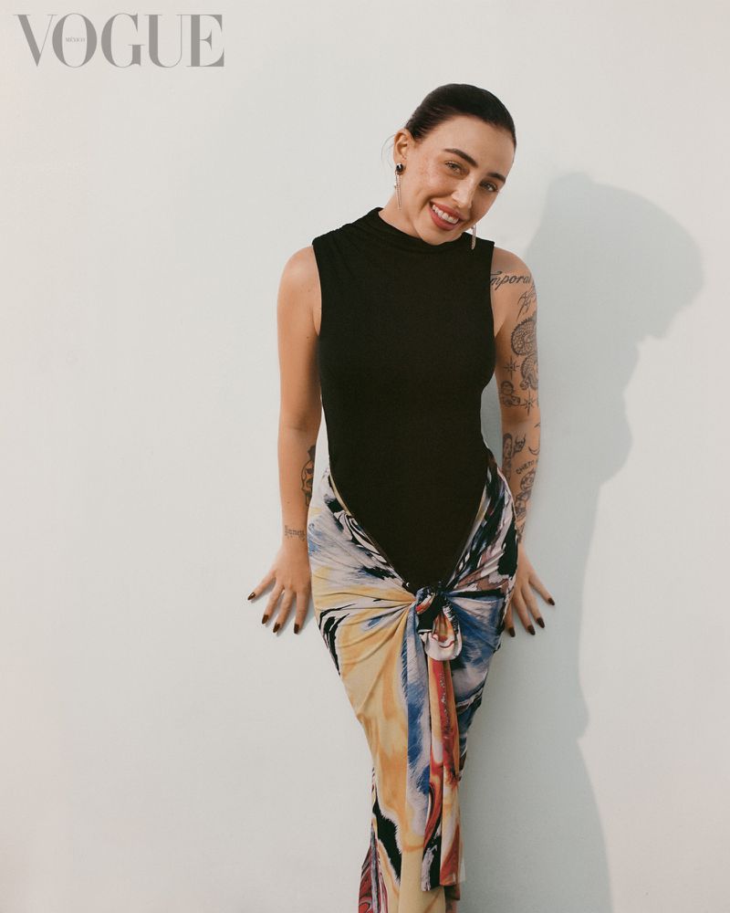 Adult female fashion model wearing a black sleeveless shirt and colorful tied skirt poses against a neutral backdrop with soft lighting.