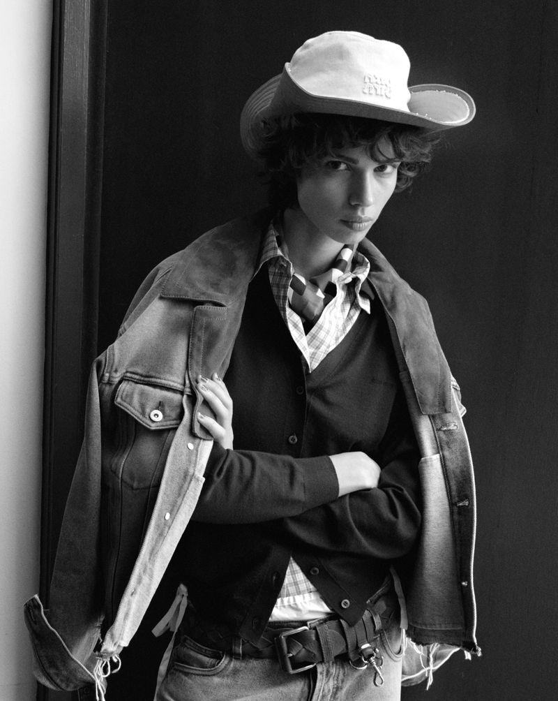 Black and white portrait of a stylish person wearing a cowboy hat, denim jacket, and cardigan, standing in front of a dark wall.
