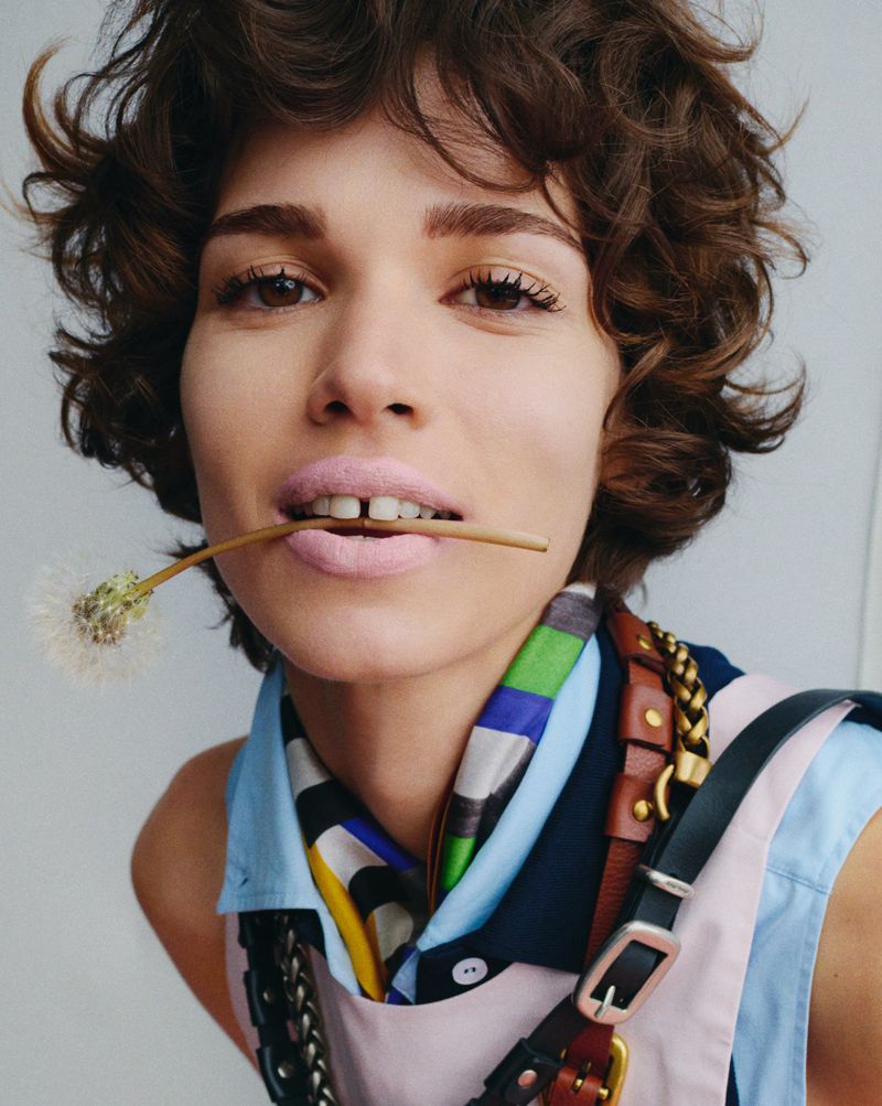 Close-up portrait of a young woman with short curly hair holding a dandelion between her lips, wearing colorful scarf and harness.