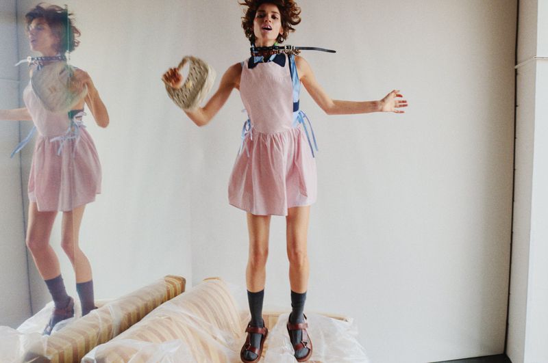 A woman in a pink dress jumps on a sofa covered in plastic in a minimalist setting, holding a basket with a duplicate of herself in the background.