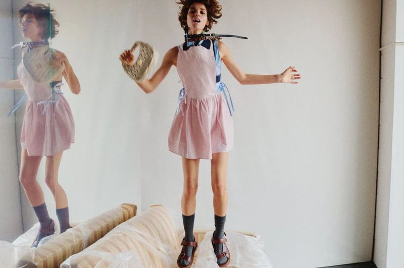 A woman in a pink dress jumps on a sofa covered in plastic in a minimalist setting, holding a basket with a duplicate of herself in the background.