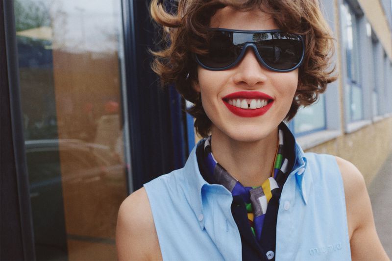 Close-up of a smiling woman with red lipstick and sunglasses in a sleeveless top, posing outdoors with a neutral urban background.