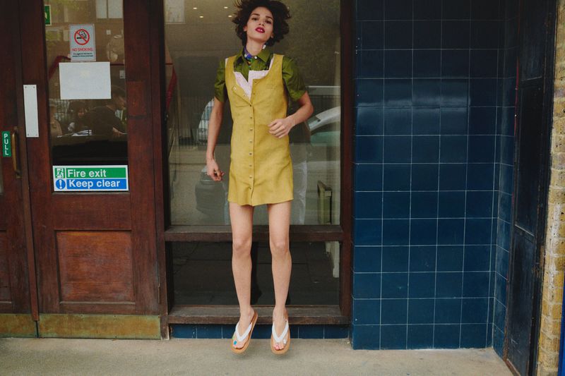 Woman jumps in a yellow mini dress and green short-sleeve shirt. Indoors near brown doors with windows and blue tiled wall.