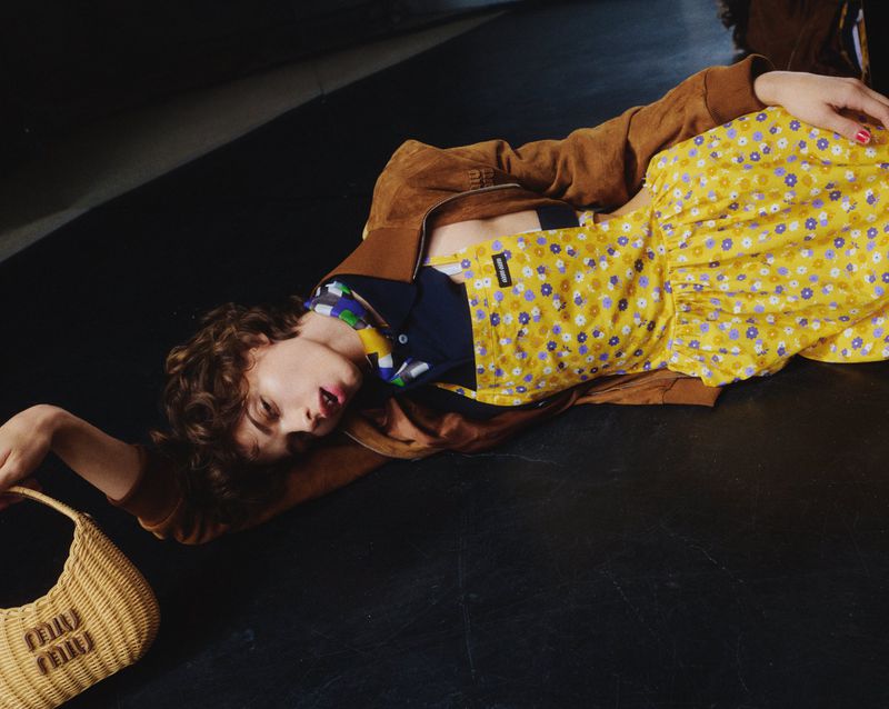 A young adult with curly brown hair lies on a dark surface, wearing a yellow floral dress, brown jacket, and holding a woven handbag.
