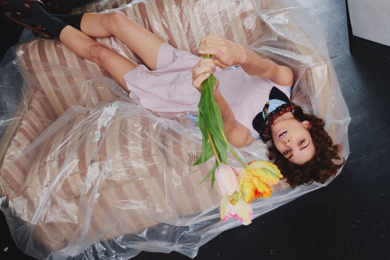 Woman lies on a vintage striped sofa covered in plastic, holding a bouquet of tulips in indoor studio setting, eye level angle.