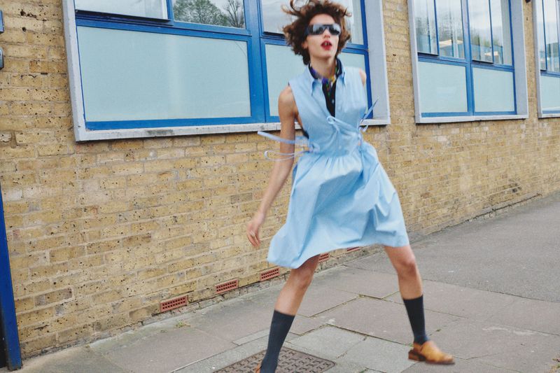 A woman with sunglasses and a light blue dress is mid-air on a city sidewalk next to a brick building with blue-framed windows.