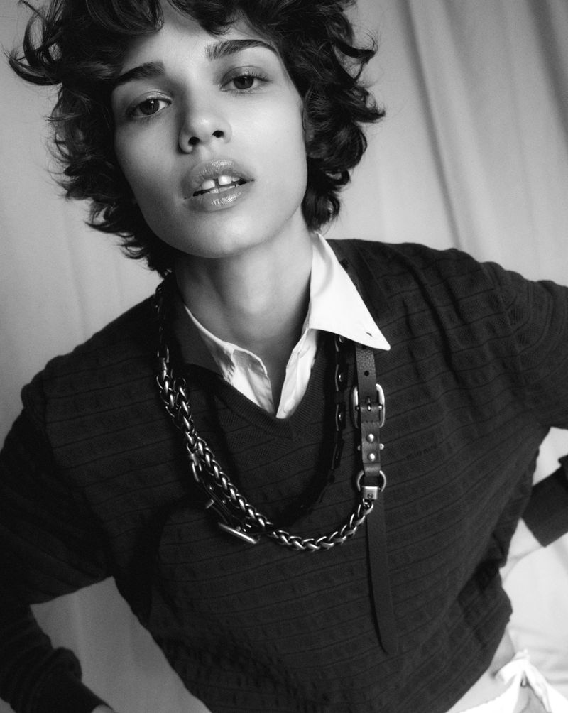 Close-up of a young adult with curly hair, wearing a dark sweater and layered collars with chains, in a monochromatic studio setting.
