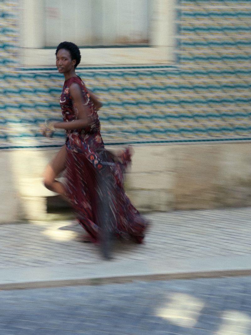 Woman in a long floral dress is running in the street. The photograph showcases motion blur and the patterned tile wall as the backdrop.