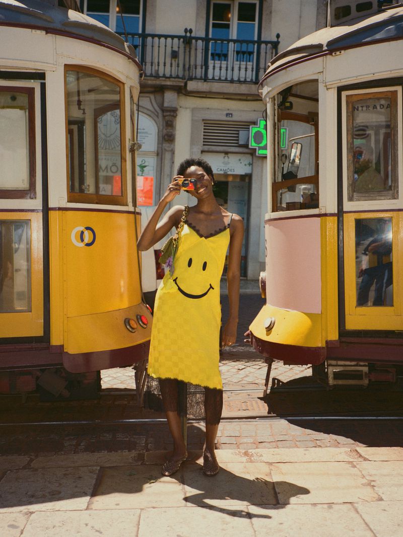 A smiling African-American woman in a yellow dress, holding sunglasses, stands between two Lisbon trams on a cobblestone street.