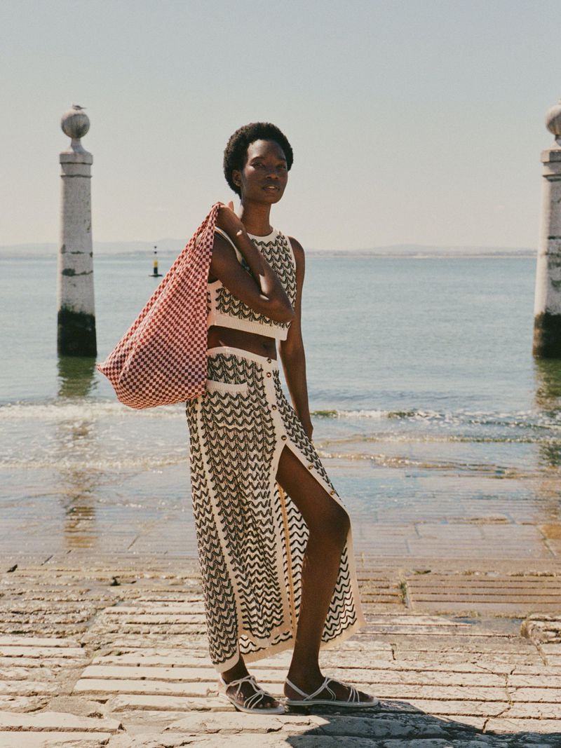 A fashionable Black woman models a knit crop-top and skirt set with red gingham tote bag, against an ocean backdrop in bright daylight.