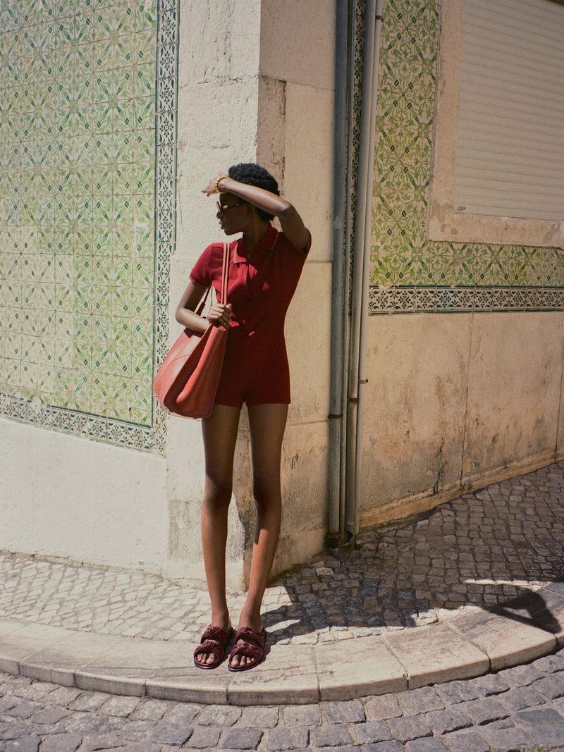 A woman in a red romper stands on a corner with patterned walls and cobblestone ground, carrying a red tote bag on a sunny day.