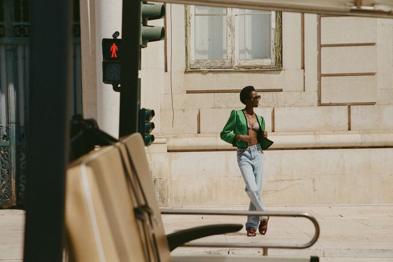 An adult female in a green jacket and jeans poses on a city sidewalk near a crosswalk, with a building in the background.