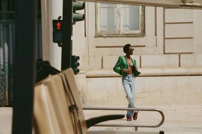 An adult female in a green jacket and jeans poses on a city sidewalk near a crosswalk, with a building in the background.