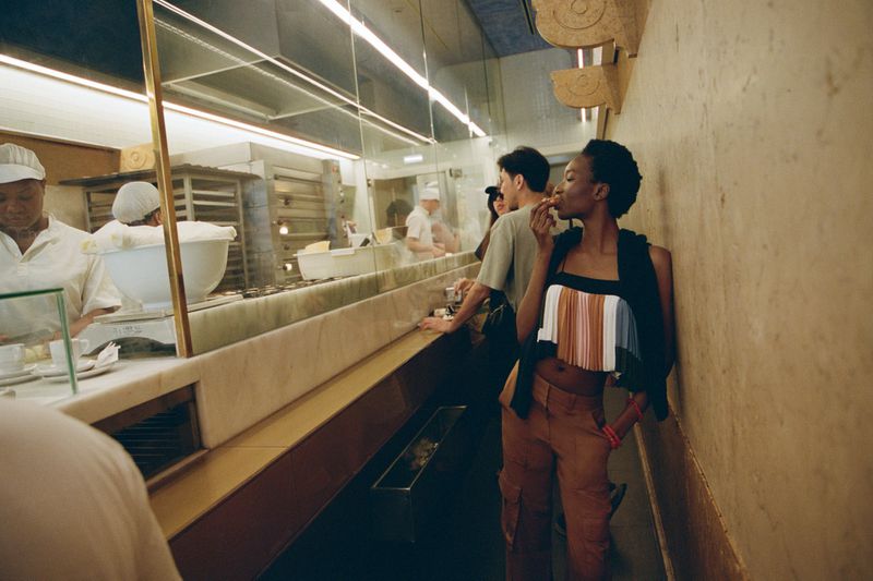 Stylish African American woman in a modern top and orange pants eating at a bakery, standing near other patrons and a marble counter.