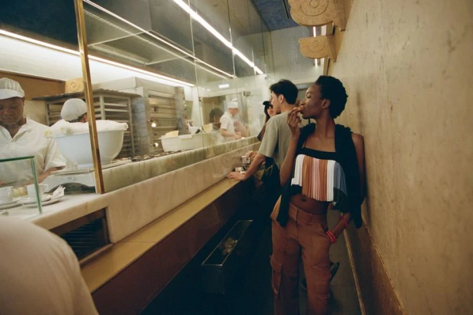 Stylish African American woman in a modern top and orange pants eating at a bakery, standing near other patrons and a marble counter.