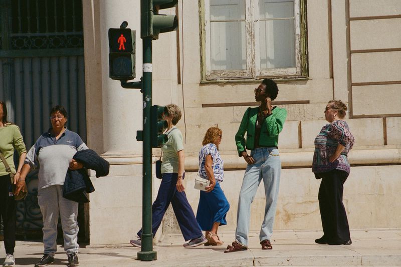 A fashionable Black woman in a green jacket and light-wash jeans stands at a crosswalk, with several people in the background in an urban setting.