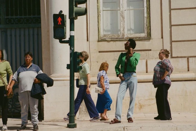 A fashionable Black woman in a green jacket and light-wash jeans stands at a crosswalk, with several people in the background in an urban setting.