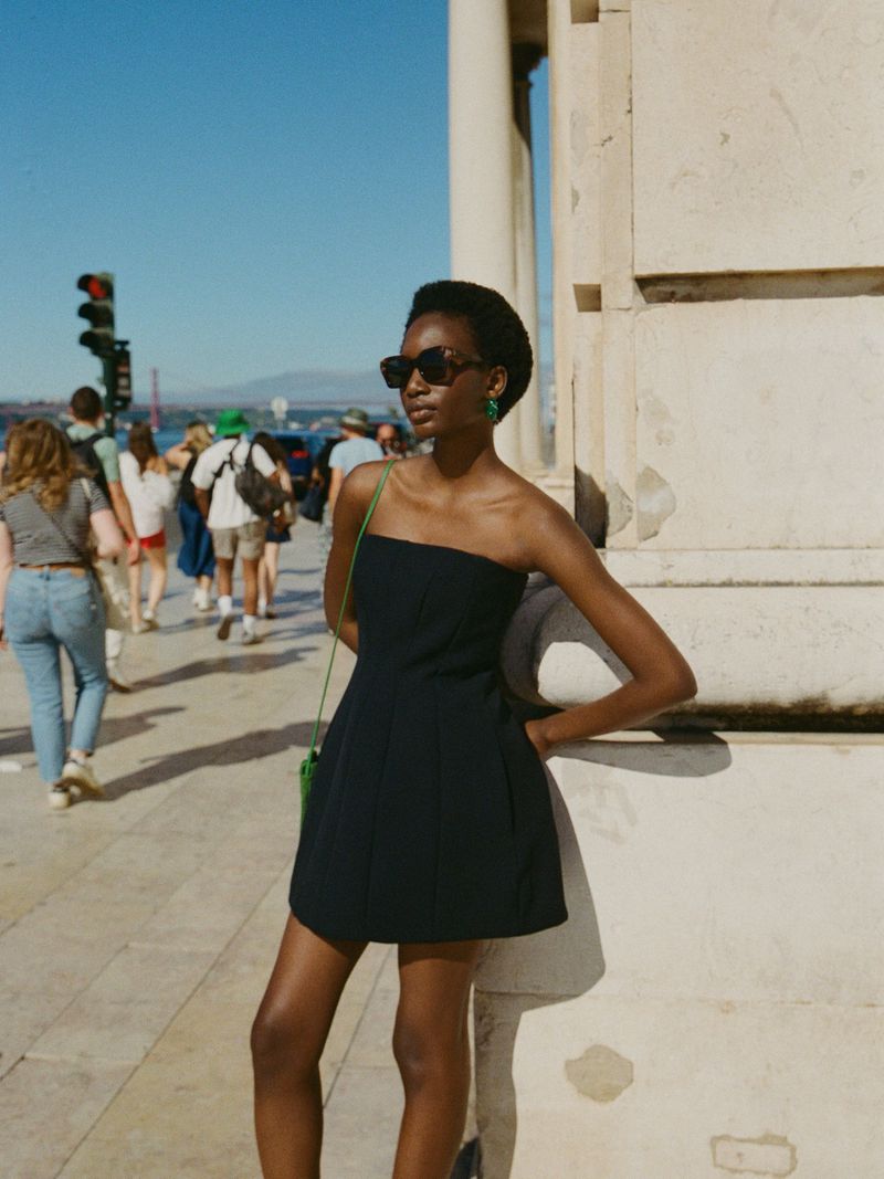 A stylish woman in a black mini dress poses on a sunny city street next to a stone wall, showcasing a modern and chic fashion ensemble.