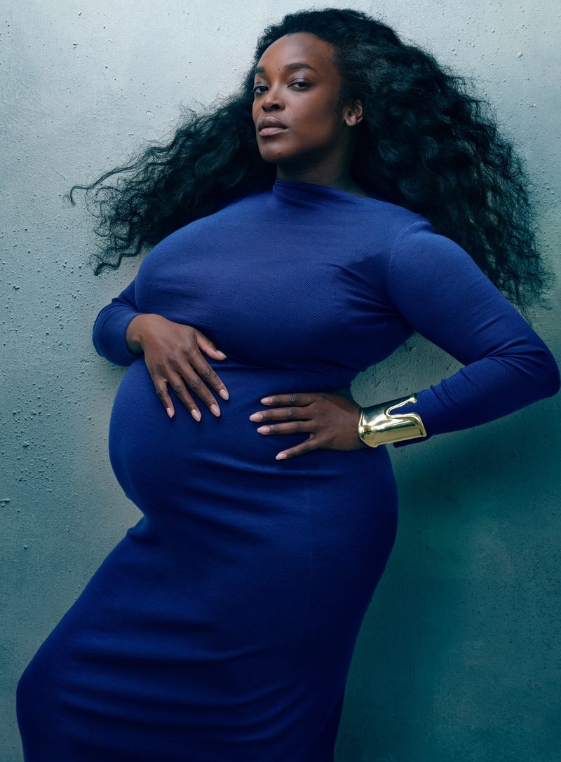 Pregnant Black woman in a blue dress poses in front of a textured gray wall, accessorized with a gold cuff bracelet on her arm.